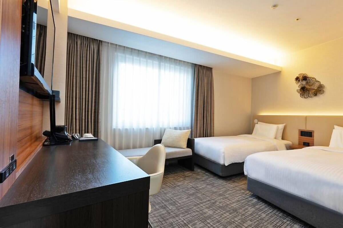 A hotel room with a desk, two beds, a chair, and a large window with curtains.