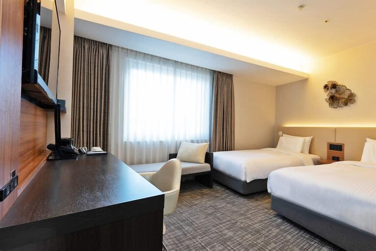 A hotel room with a desk, two beds, a chair, and a large window with curtains.