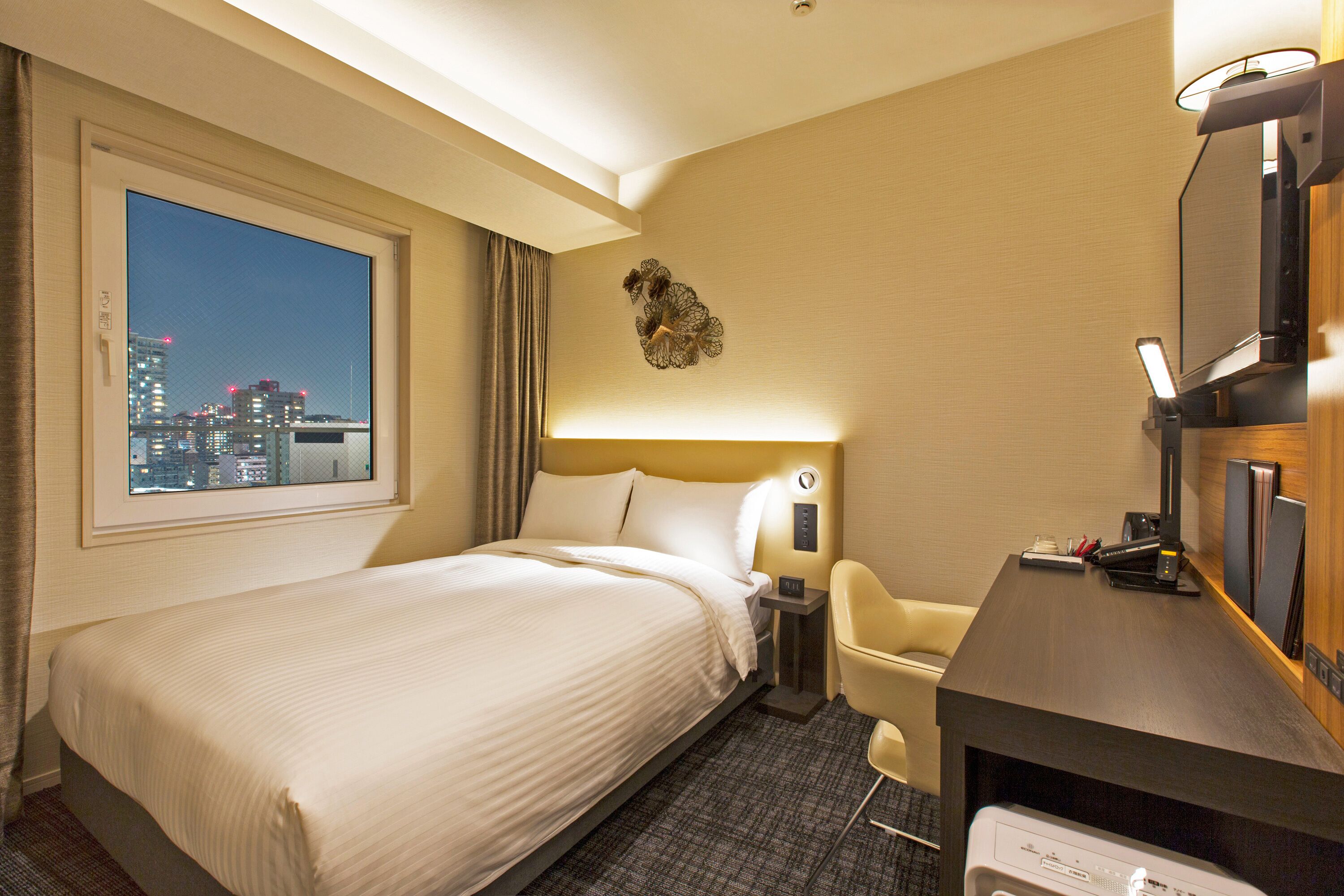 Ein Hotelzimmer mit einem großen Bett, einem Schreibtisch mit Computer, einem Sessel und einer Lampe, mit Blick auf die beleuchtete Stadt bei Nacht.