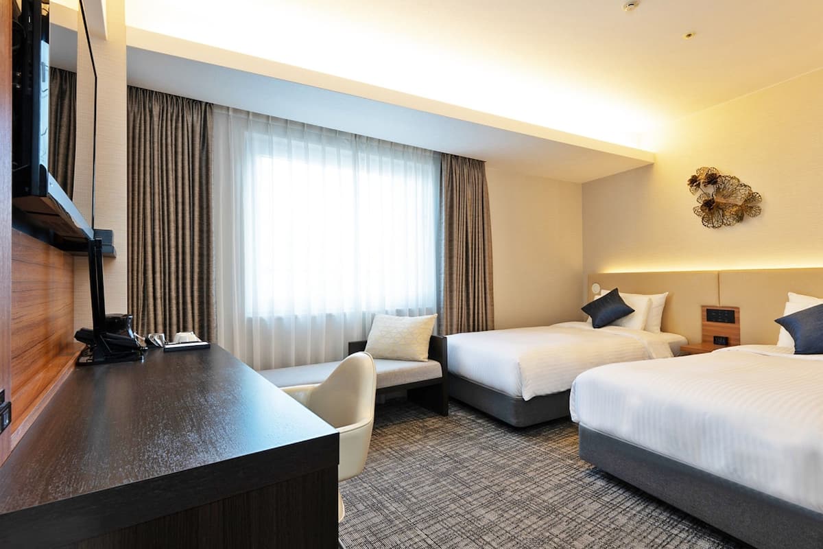 A hotel room with two beds, a desk, a chair, and a large window with curtains.