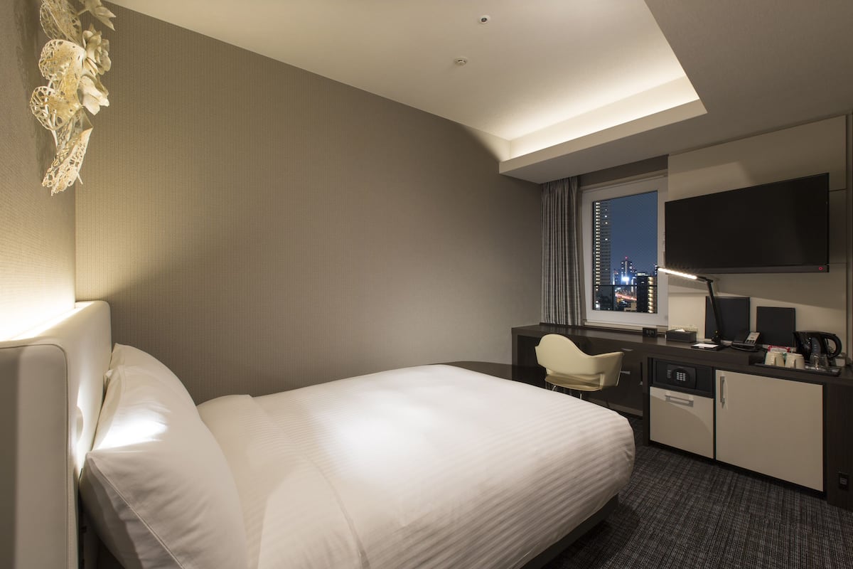 A hotel room with a large bed, a desk with a chair, a television, and a window with a city view.