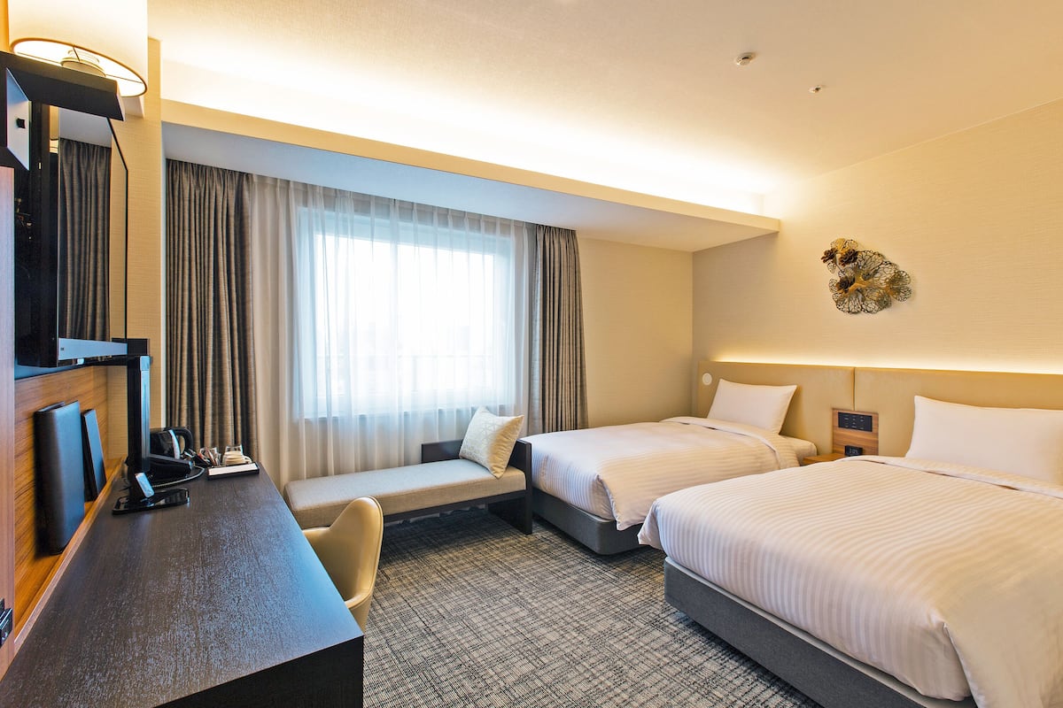 A hotel room with two beds, a desk, a chair, and a television.