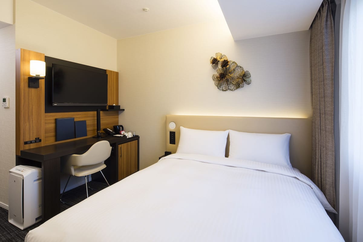 A hotel room with a bed, a desk with a chair, a flat-screen TV, a small desk lamp, and a decorative wall piece.