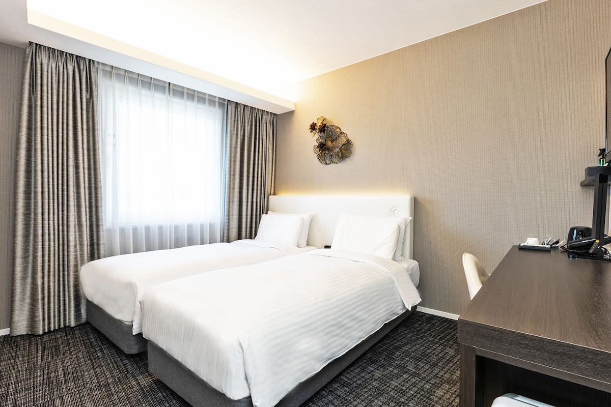 A hotel room with a double bed, a desk, a chair, a small table, a telephone, and a decorative wall piece.