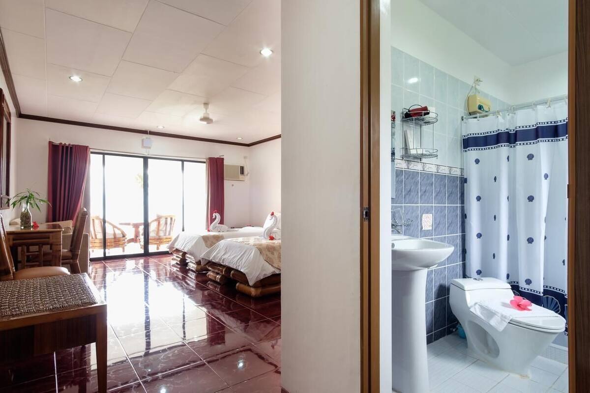 Habitación de hotel con cama, una mesa de comedor con sillas, un ventanal grande y un baño con cortina de ducha.