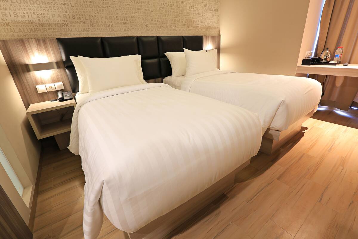 Habitación de hotel con dos camas, piso de madera, cabecera con papel tapiz con el nombre de una ciudad, mesita de noche con lámpara y una pequeña mesa con una botella y vasos.