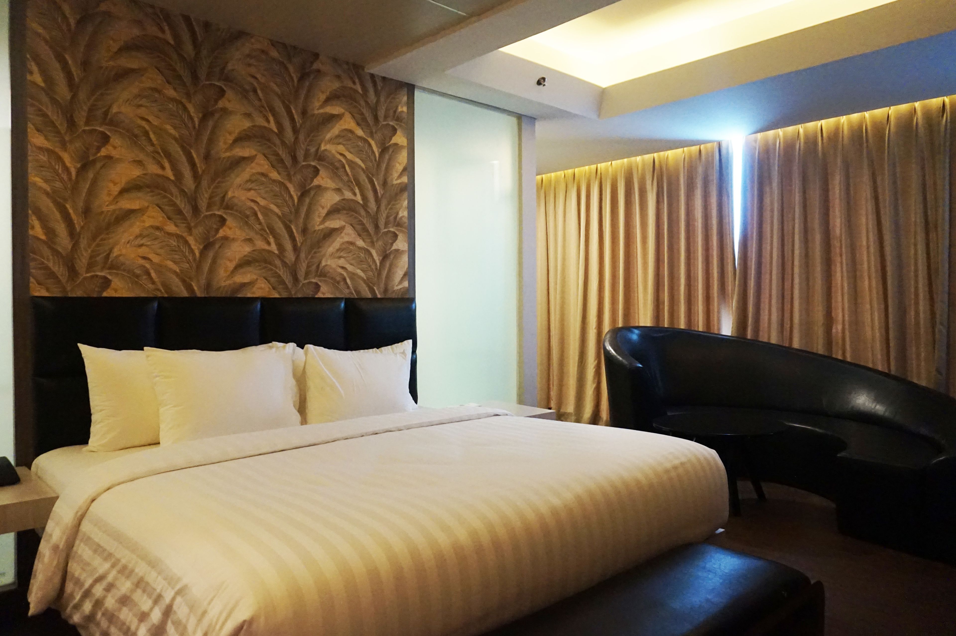 Una habitación de hotel con una cama grande, un cabecero con diseño de hojas, un sillón de cuero negro y una ventana con cortinas.