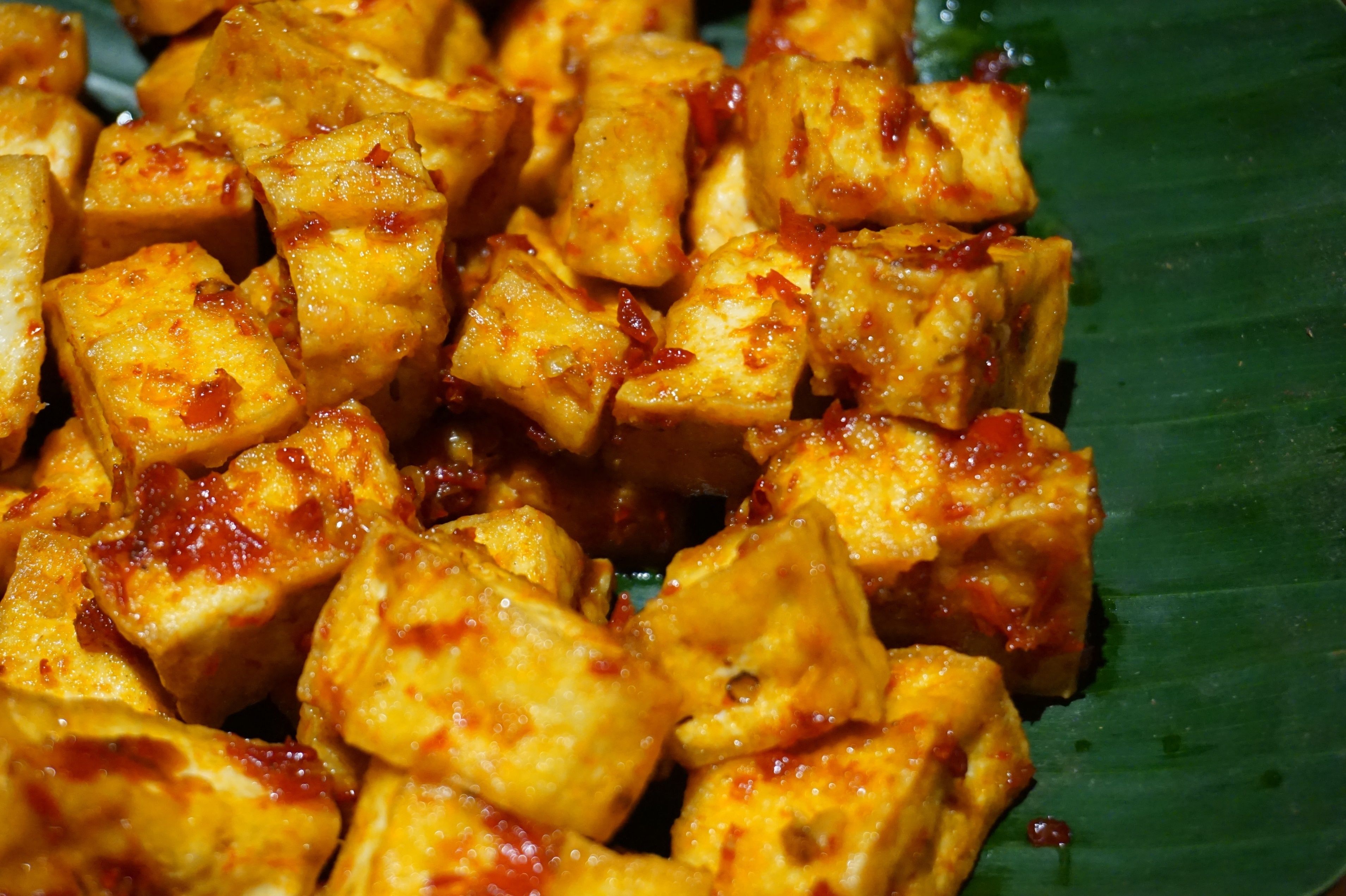 A close-up of cubed tofu with a reddish sauce, served on a green leaf.