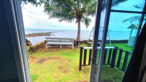 Vue depuis une pièce dont la porte est en verre, donnant sur un espace verdoyant avec un banc et un palmier, et d’où l’on aperçoit un littoral rocheux et l’océan.