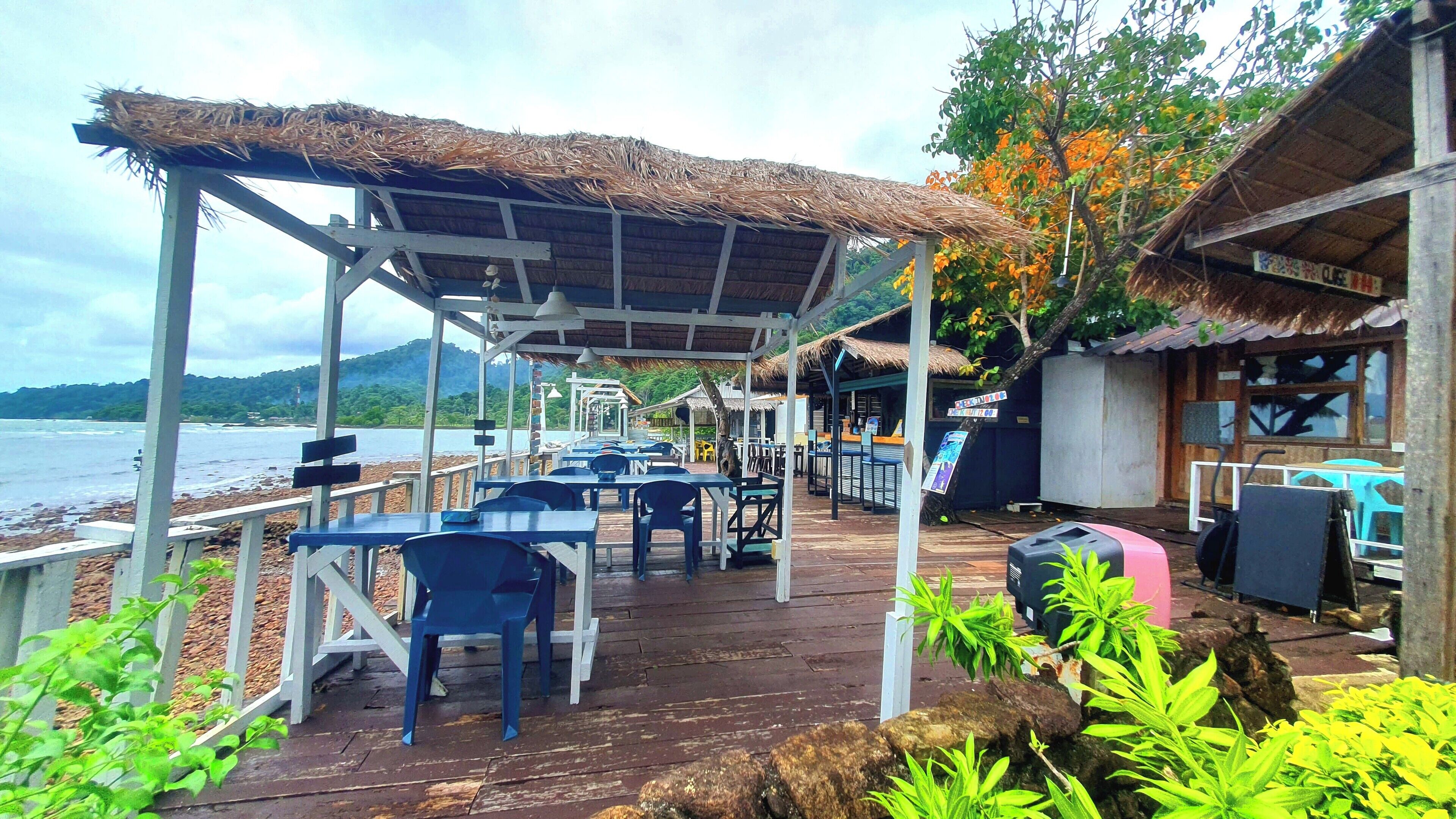 An outdoor seating area with tables and chairs, a thatched roof, and a view of the sea.