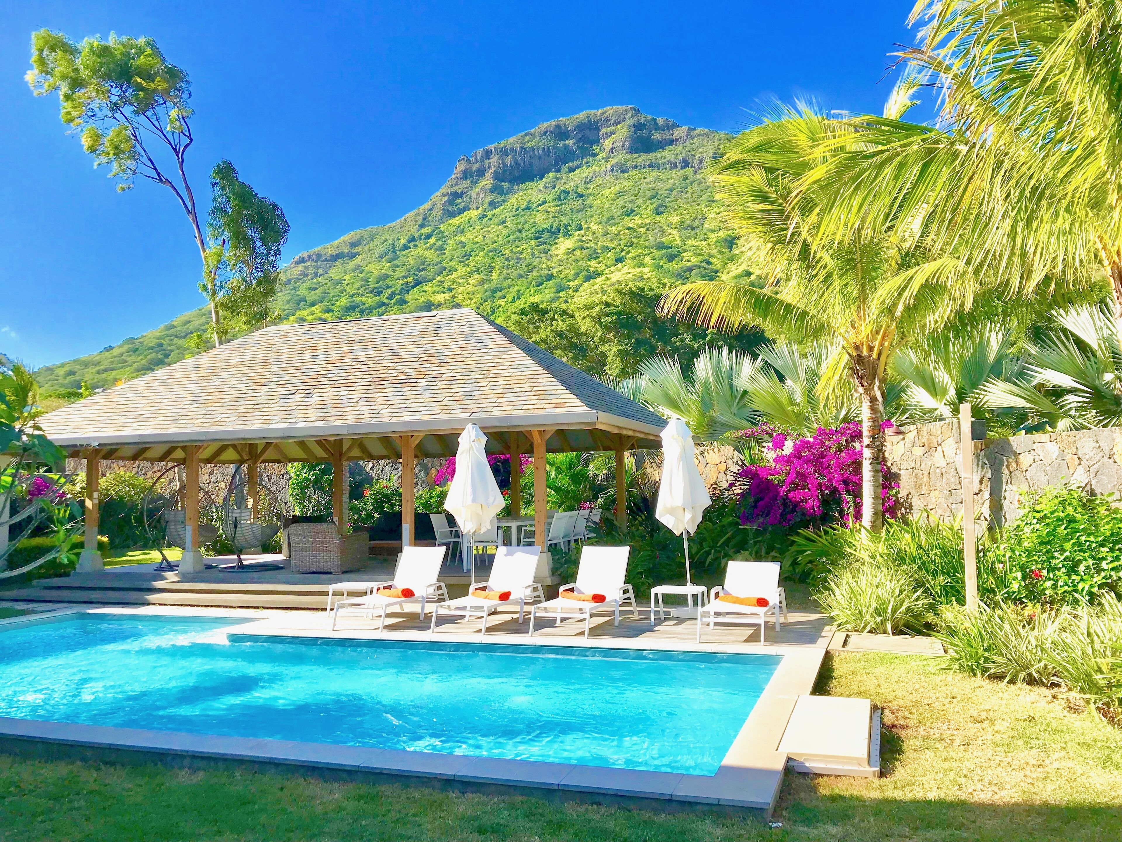 A pool area with lounge chairs and a gazebo, surrounded by lush greenery and a mountain backdrop.