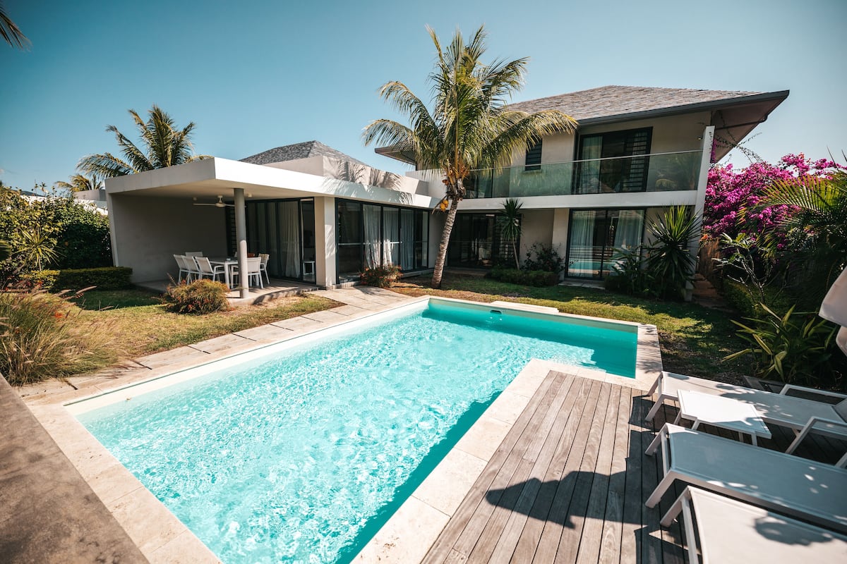 Un área de piscina moderna con terraza de madera, sillas de descanso y una casa de dos pisos con ventanas amplias y un porche cubierto.