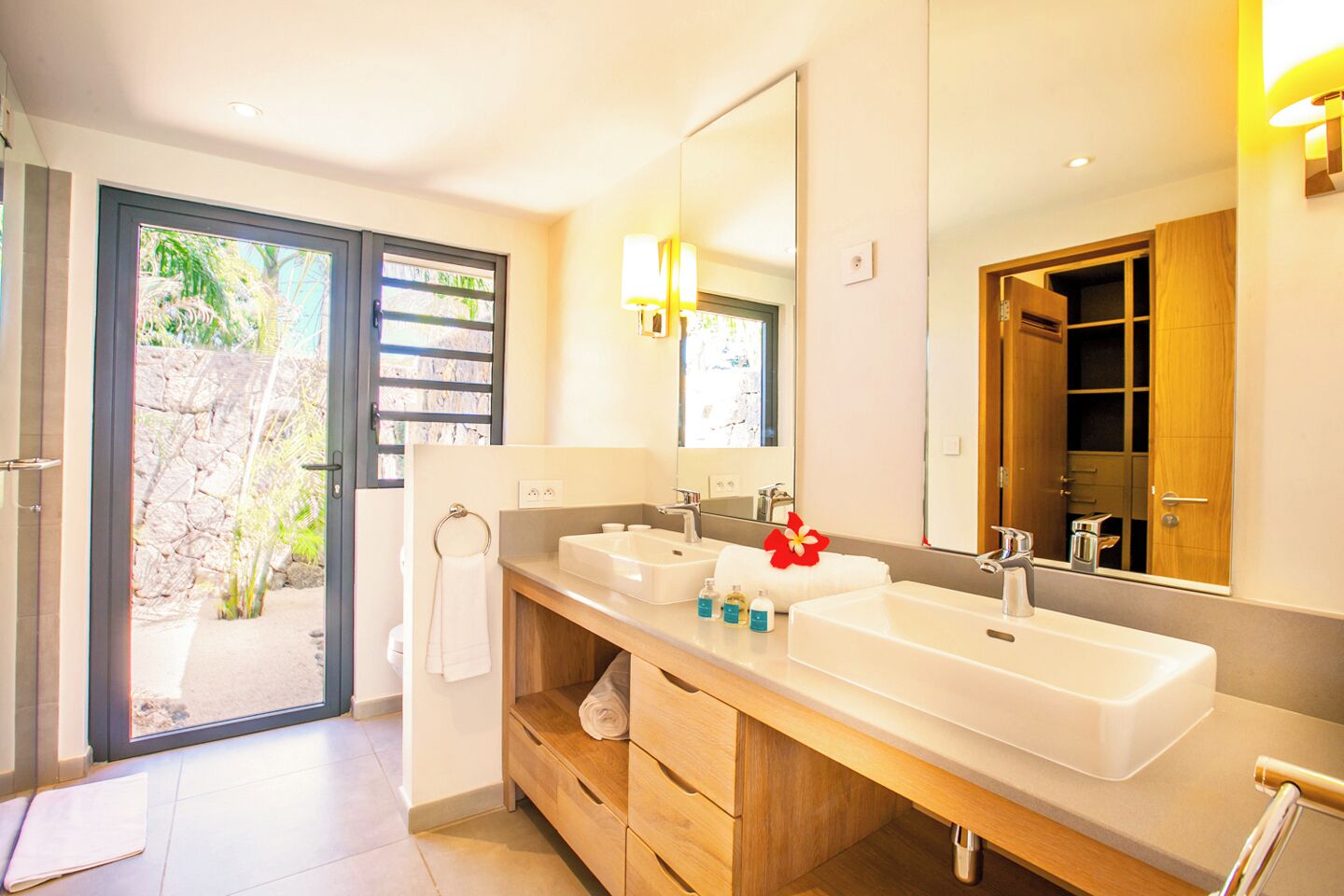 Ein modernes Badezimmer mit zwei Waschbecken, einem großen Spiegel und Blick auf einen Garten durch die Tür.