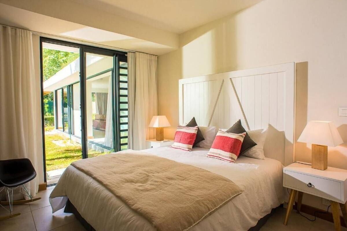 Un dormitorio con cama, dos mesitas de noche con lámparas, una silla y un ventanal corredero que da a un área exterior.