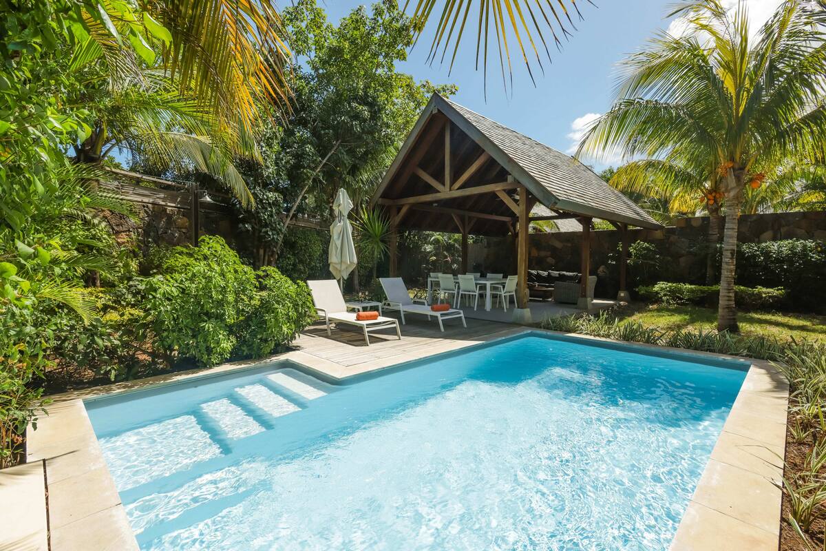 Piscina con sillones de descanso y un área de estar cubierta, rodeada de exuberante vegetación.