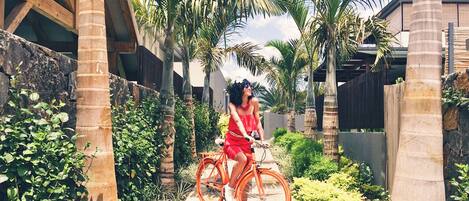Una persona pedalea una bicicleta sobre un sendero de concreto rodeado de follaje y palmeras.