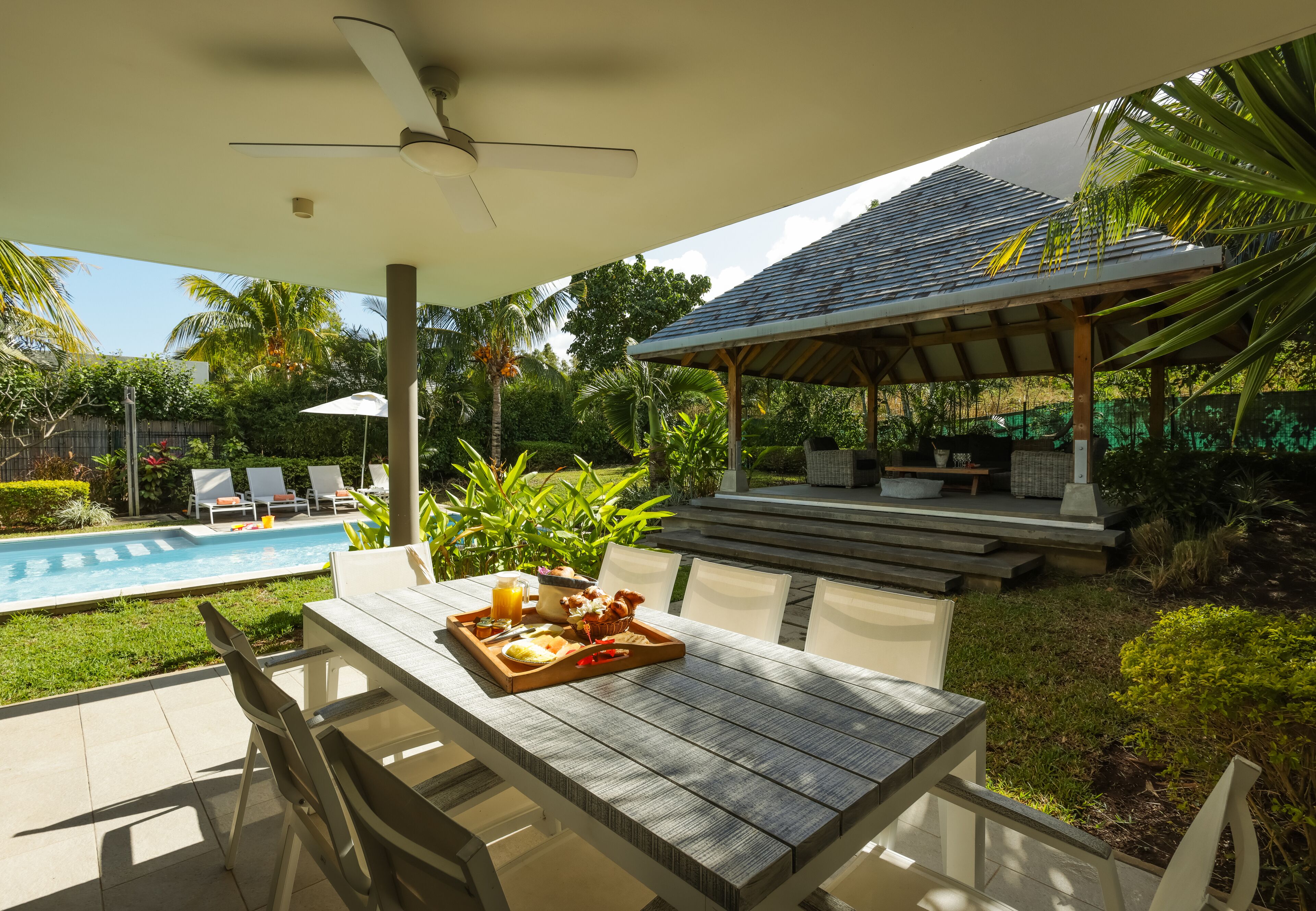 A covered outdoor dining area with a table set for breakfast, overlooking a swimming pool and lush greenery.