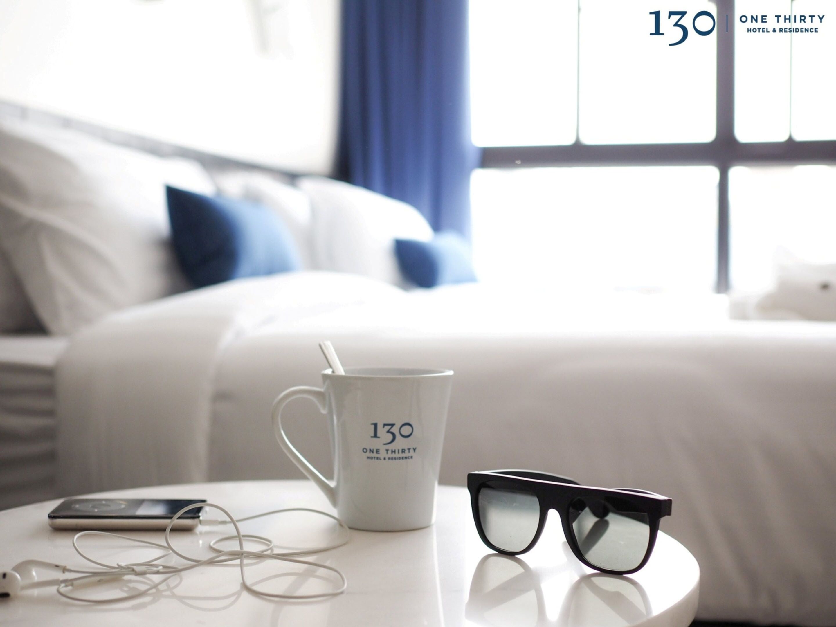 A white mug with "130 ONE THIRTY HOTEL & RESIDENCE" text, black sunglasses, and white earphones on a white table in a hotel room with a bed and blue curtains in the background.