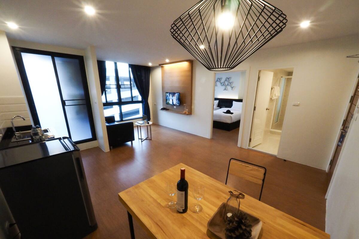 Une chambre d’hôtel moderne comprenant une table à manger en bois, un coin cuisine, un espace de vie avec un téléviseur et une chambre à coucher avec un lit et une table de chevet.
