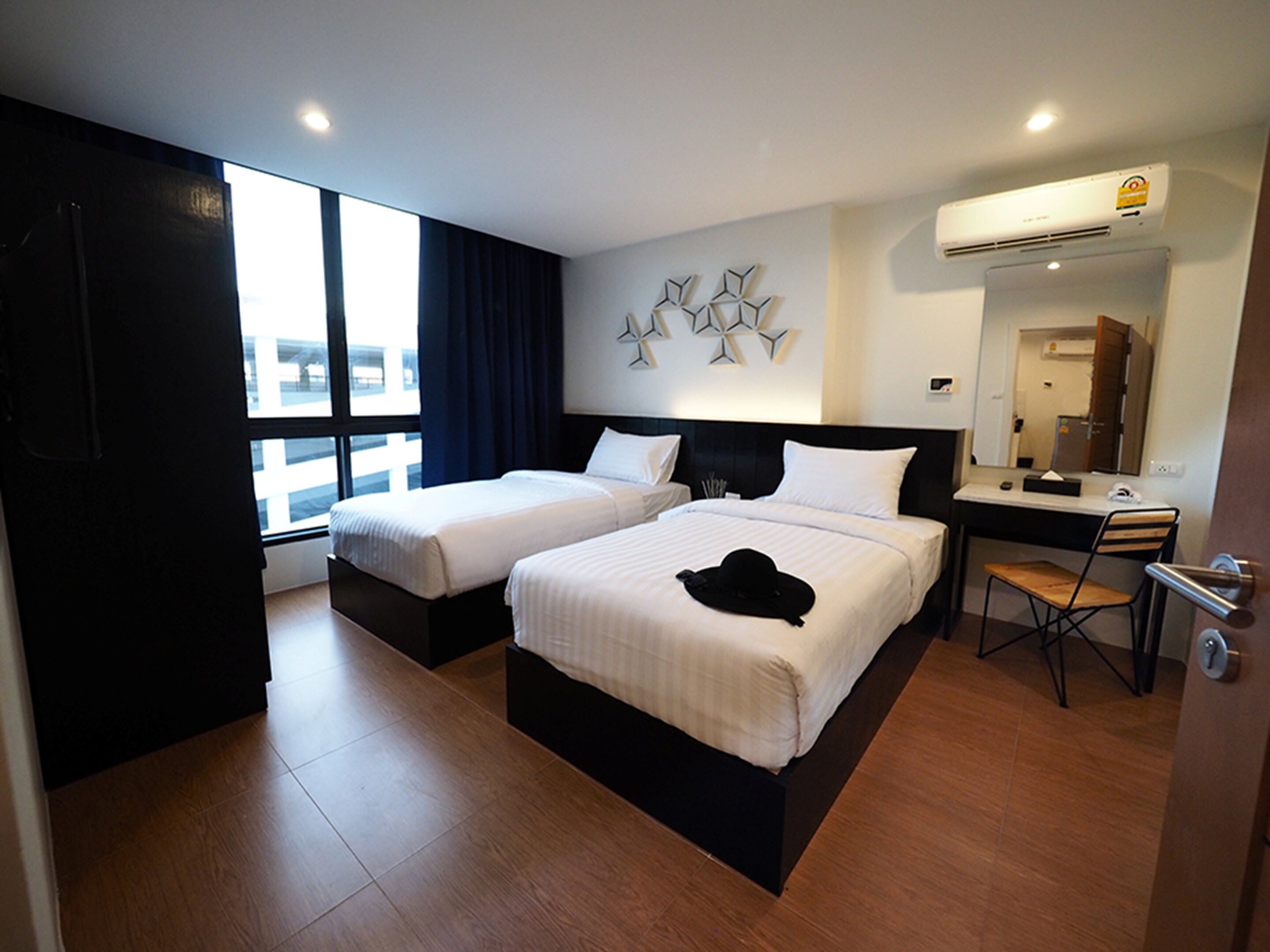 Habitación de hotel con dos camas, un escritorio y una silla. Tiene una ventana con cortinas, un aire acondicionado y un espejo.