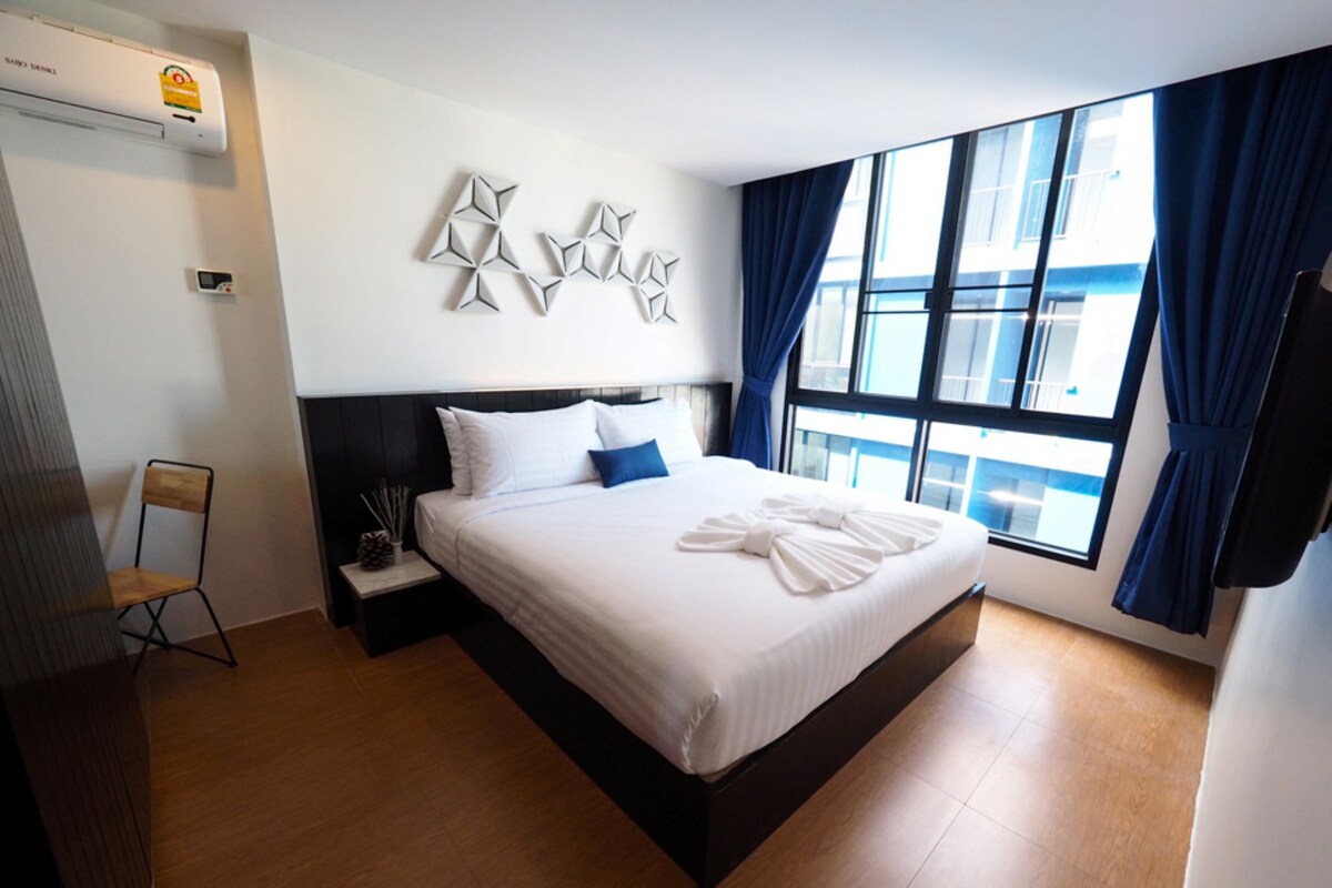 Ein Hotelzimmer mit einem großen Bett, einem Nachttisch, einem Sessel, einem Fenster mit Vorhängen und einer Wand-Klimaanlage.