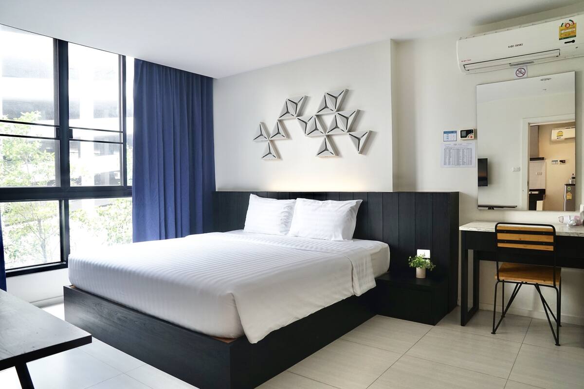 Une chambre d’hôtel moderne avec un grand lit, un bureau et une chaise. On y trouve une fenêtre avec des rideaux, un climatiseur fixé au mur et un élément décoratif mural.