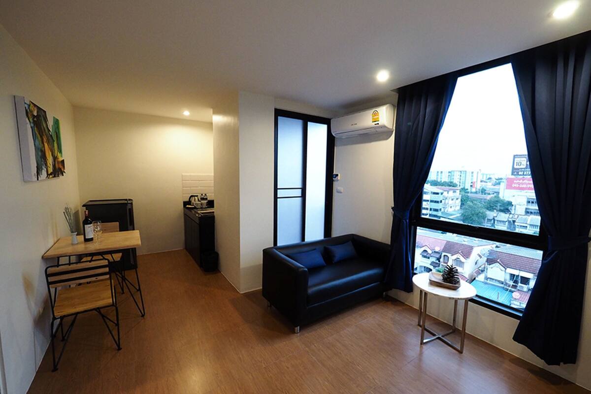 Un espace de vie compact comprenant un canapé, une petite table, un coin cuisine et une grande fenêtre avec des rideaux.