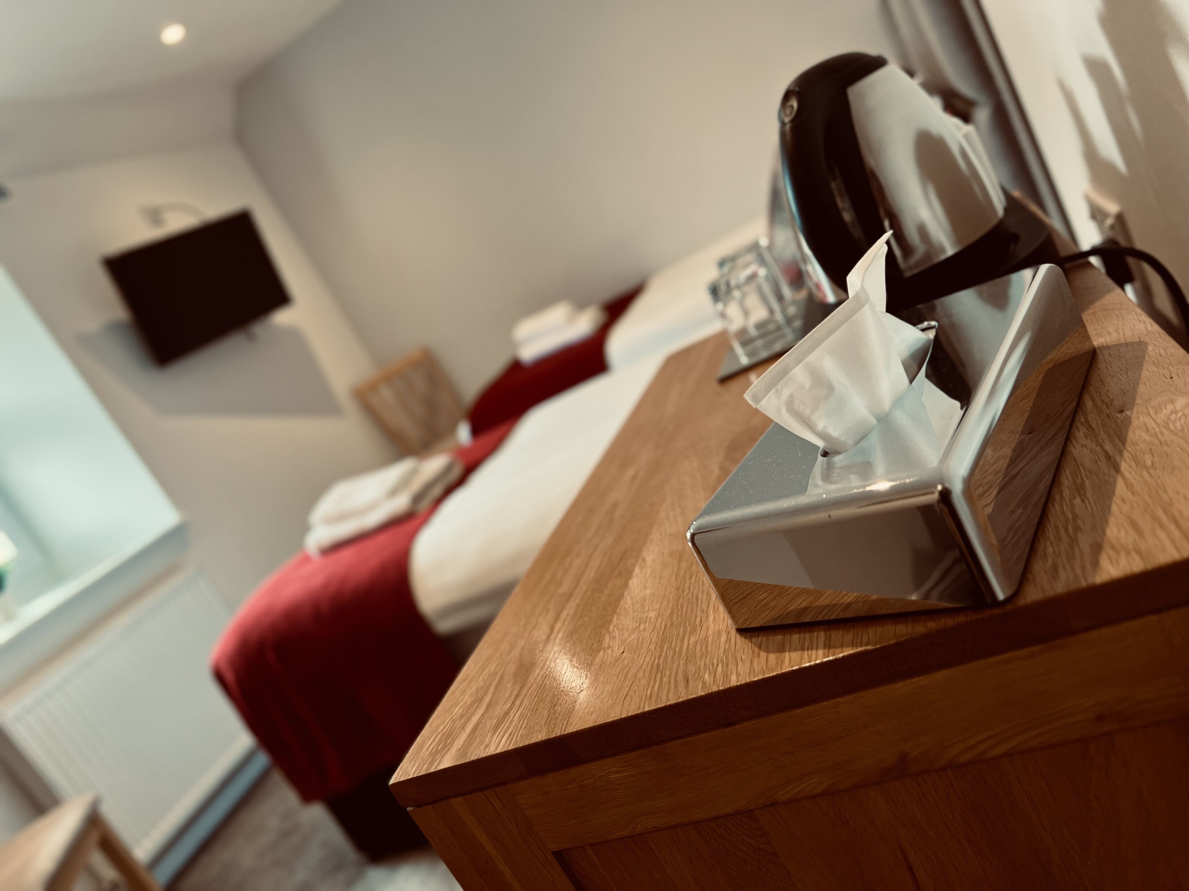 A hotel room with a wooden desk, a bedside table with a tissue box, a red bedspread, and a mounted television on the wall.