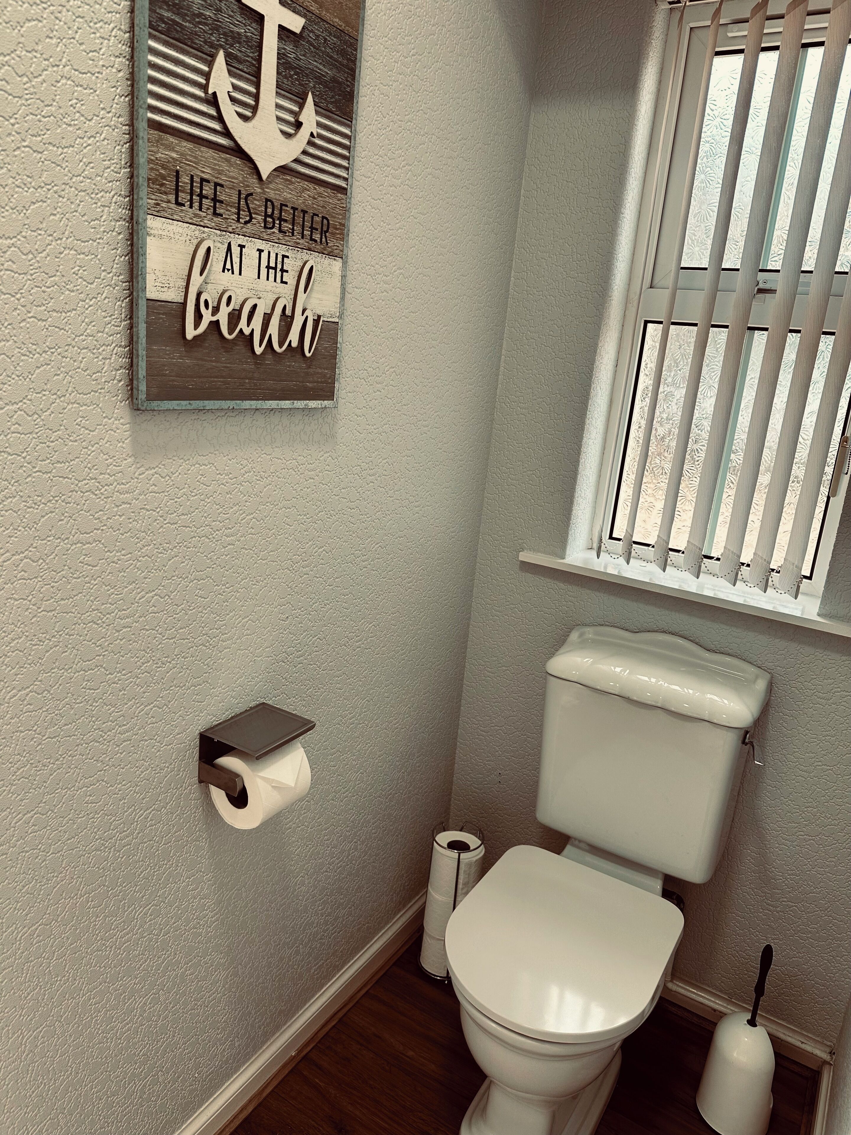 A bathroom with a toilet, a roll of toilet paper, a window with blinds, and a decorative sign on the wall.
