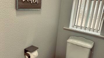 A bathroom with a toilet, a roll of toilet paper, a window with blinds, and a decorative sign on the wall.