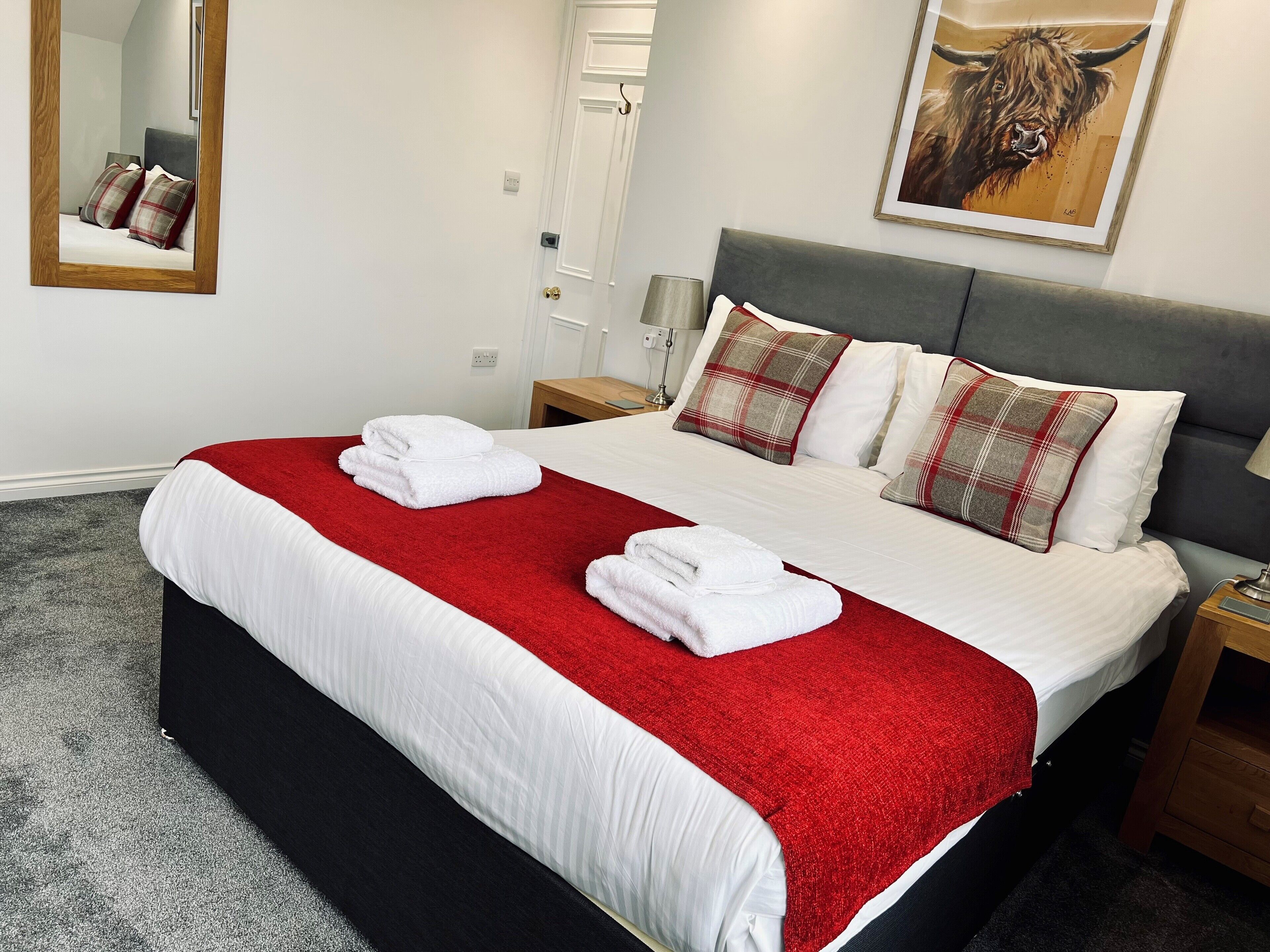 A neatly made bed with white linens and a red blanket, two pillows with a plaid pattern, a wooden nightstand, a lamp, and a painting of a bull on the wall.