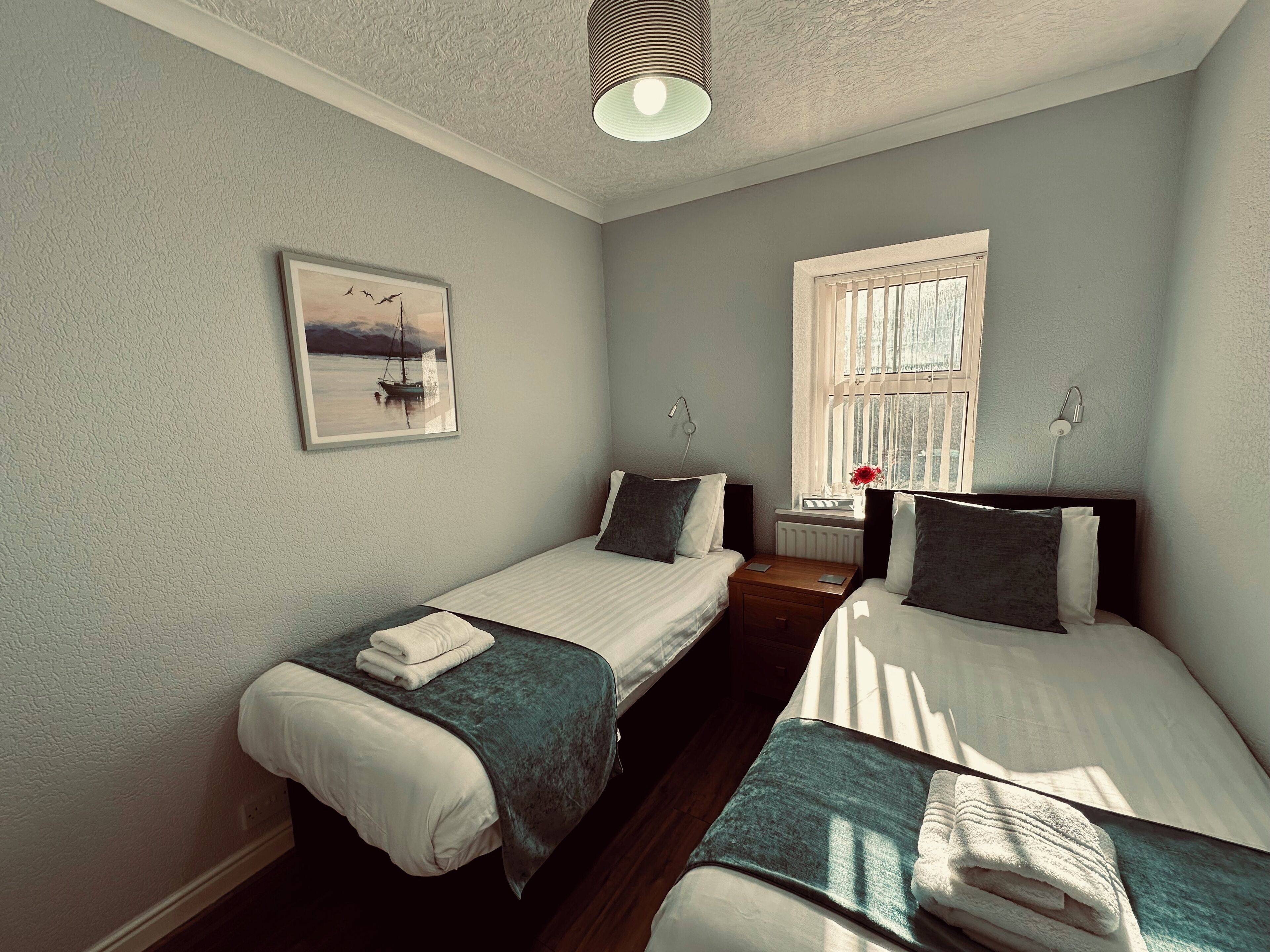 A bedroom with two single beds, a nightstand, a lamp, a framed picture, and a window with curtains.