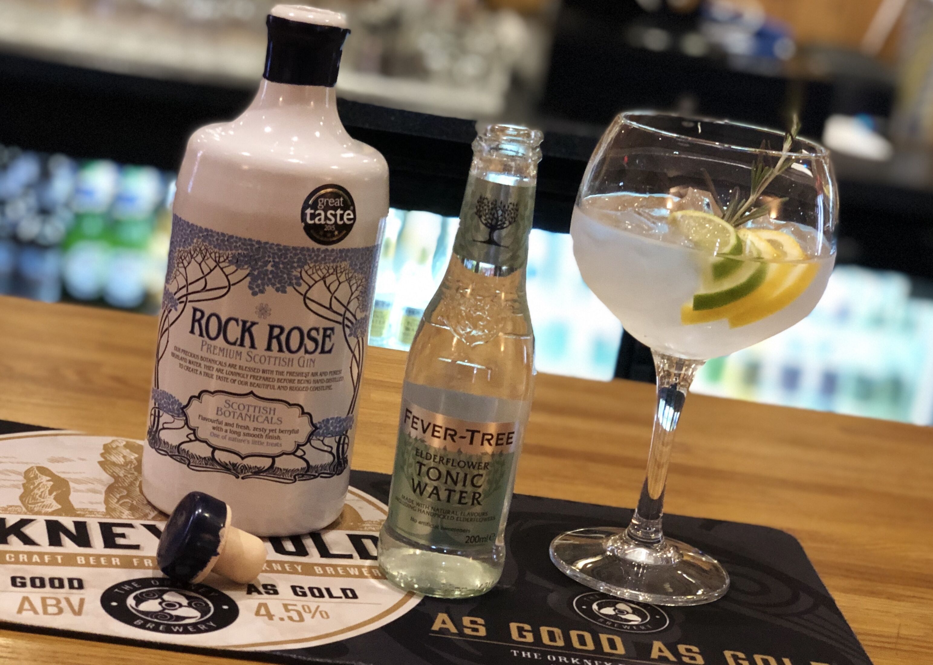 Una botella de ginebra Rock Rose, una botella de agua tónica y un vaso de ginebra tónica sobre un tapete de bar.