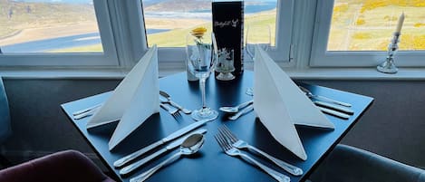 A dining table set for two with a view of a coastal landscape through large windows.