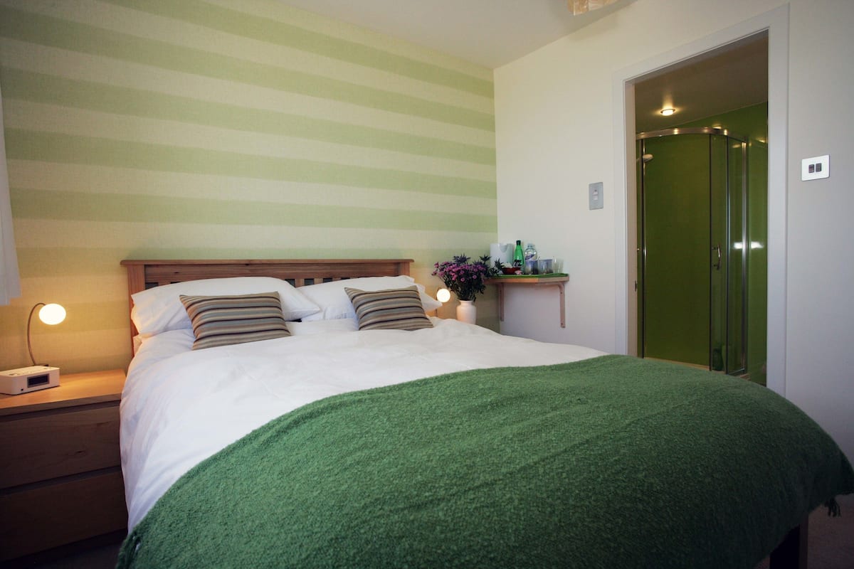 Una cama bien tendida con un edredón verde, una lámpara de noche y una mesita de noche con un radio.