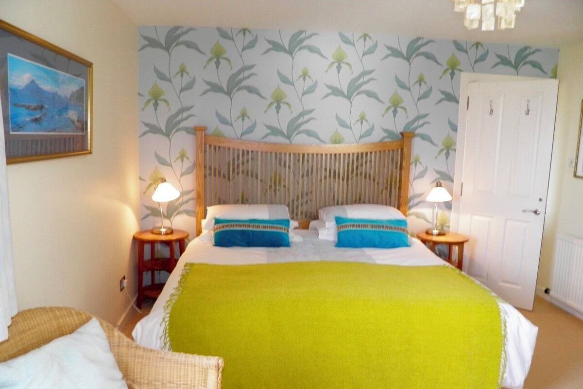 Un dormitorio con una cama, dos mesitas de noche, una mesita auxiliar, una puerta y una pared con estampado floral.