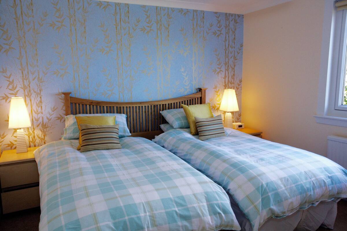 Una cama doble con cubrecama a cuadros, dos mesitas de noche con lámparas y una pared con estampado de bambú.