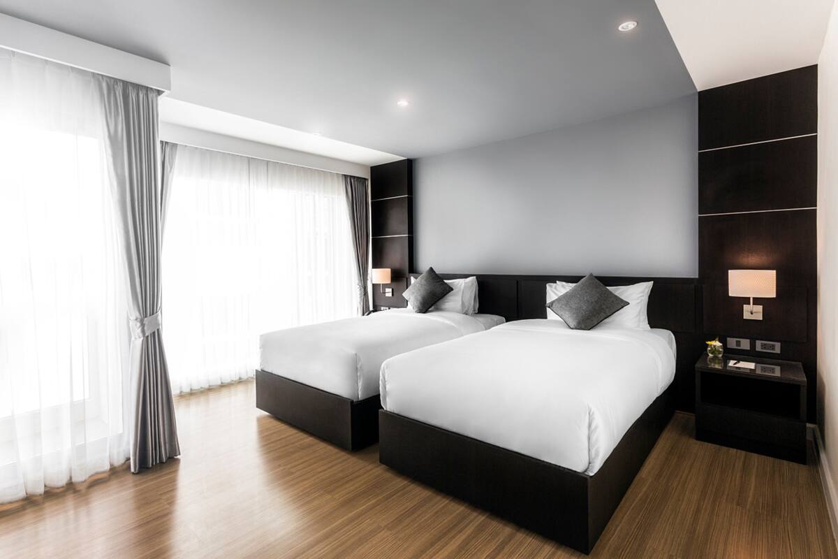 A modern hotel room with two beds, a large window with sheer curtains, a dark wood floor, and a wall-mounted nightstand with a lamp.