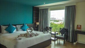 A hotel room with a large bed, two blue pillows, a desk with a chair, and a view of a cityscape through the window.