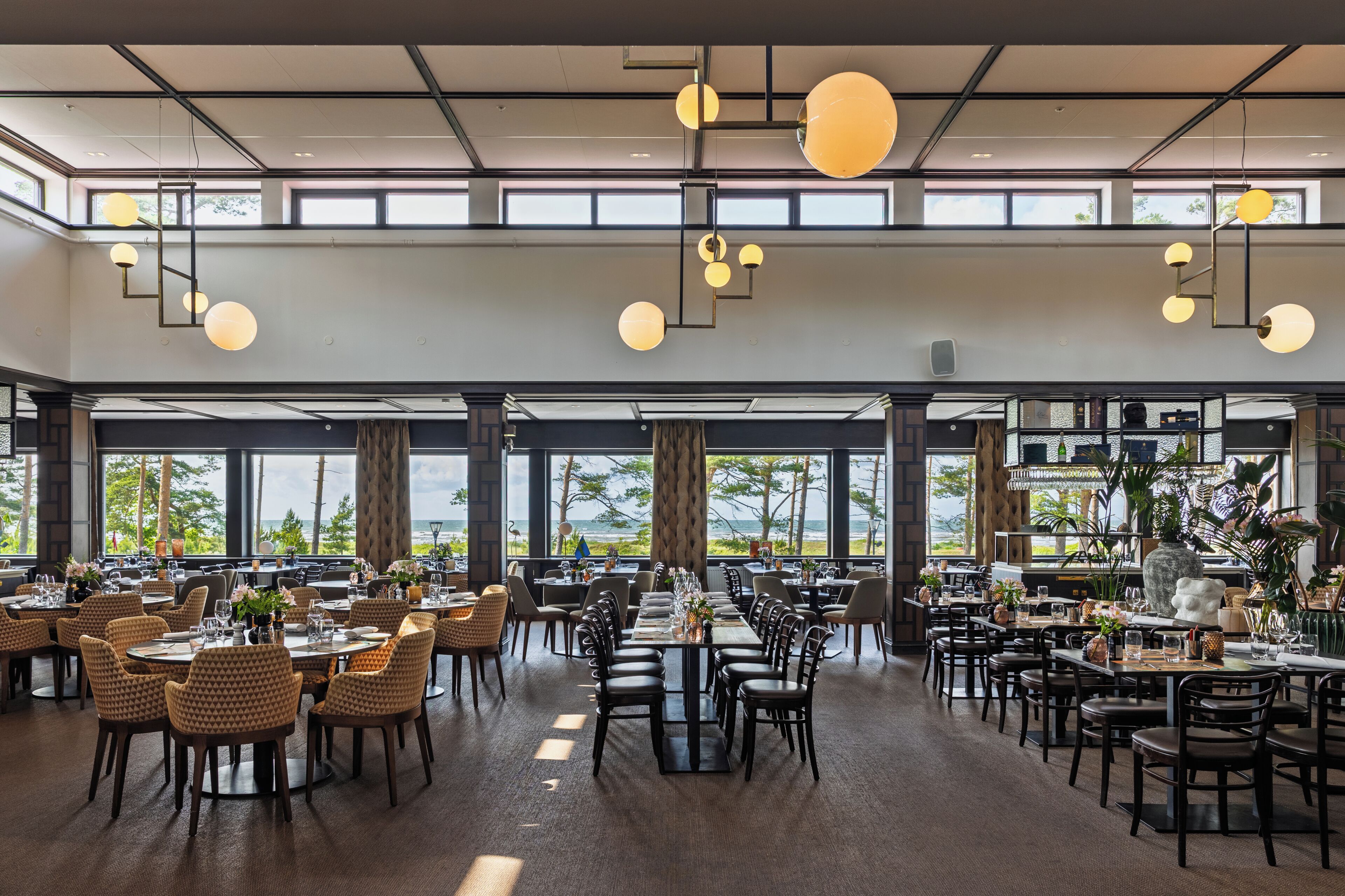Un comedor espacioso con varias mesas y sillas, ventanas amplias con vistas y lámparas colgantes esféricas.