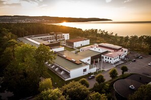 An aerial view of a modern building complex with multiple buildings, a parking area, and a view of a lake and forested hills in the background.