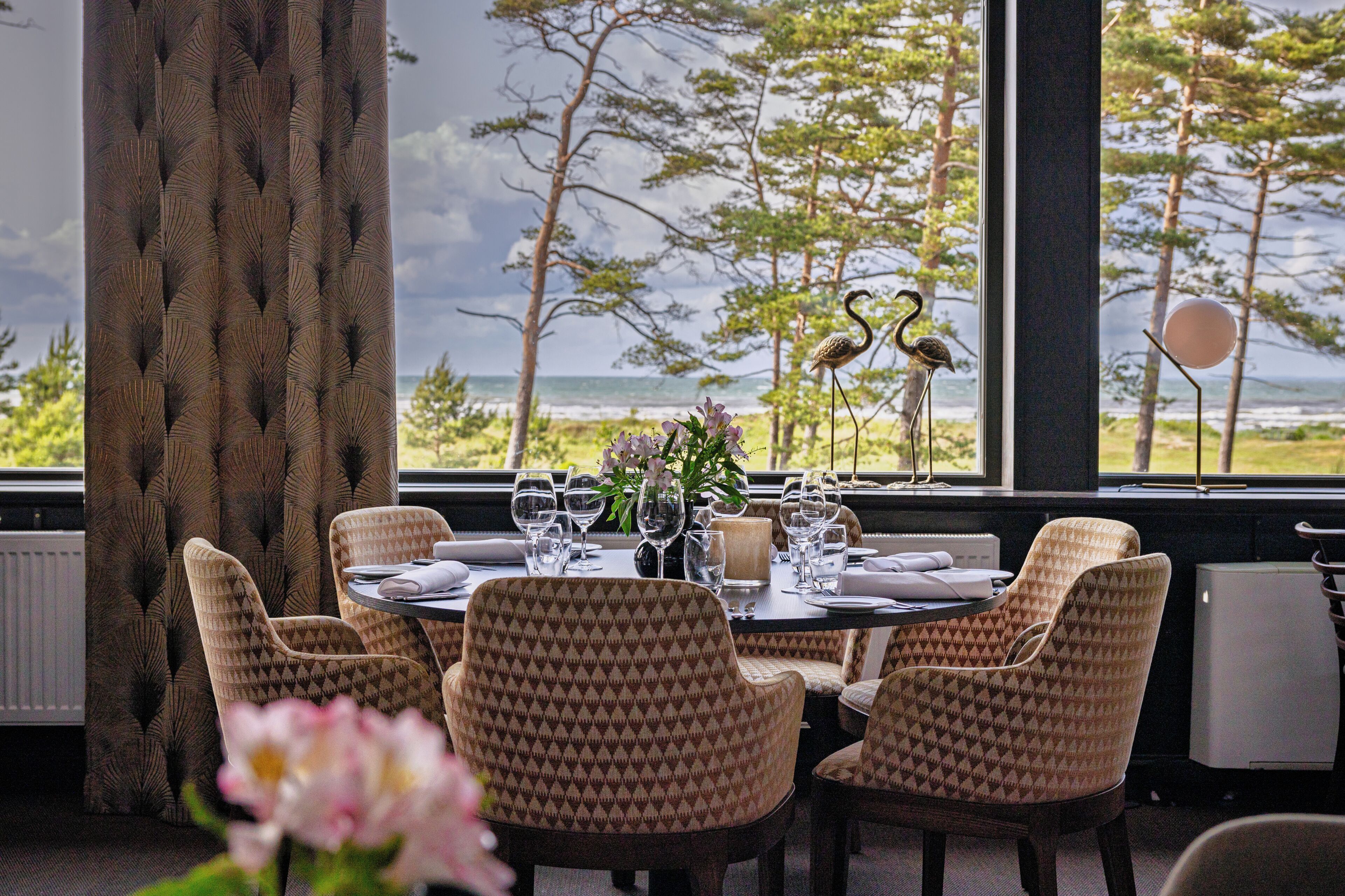 Uma área de jantar com uma mesa posta para quatro pessoas, cadeiras, um vaso com flores e vista para o oceano através de grandes janelas.