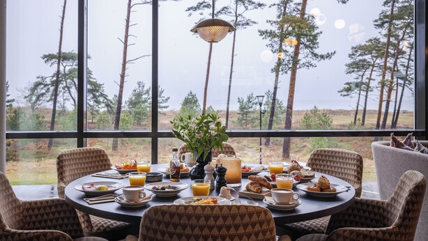 Mesa de comedor dispuesta con alimentos para el desayuno, rodeada de sillas estampadas, con vistas a altos árboles a través de amplios ventanales.