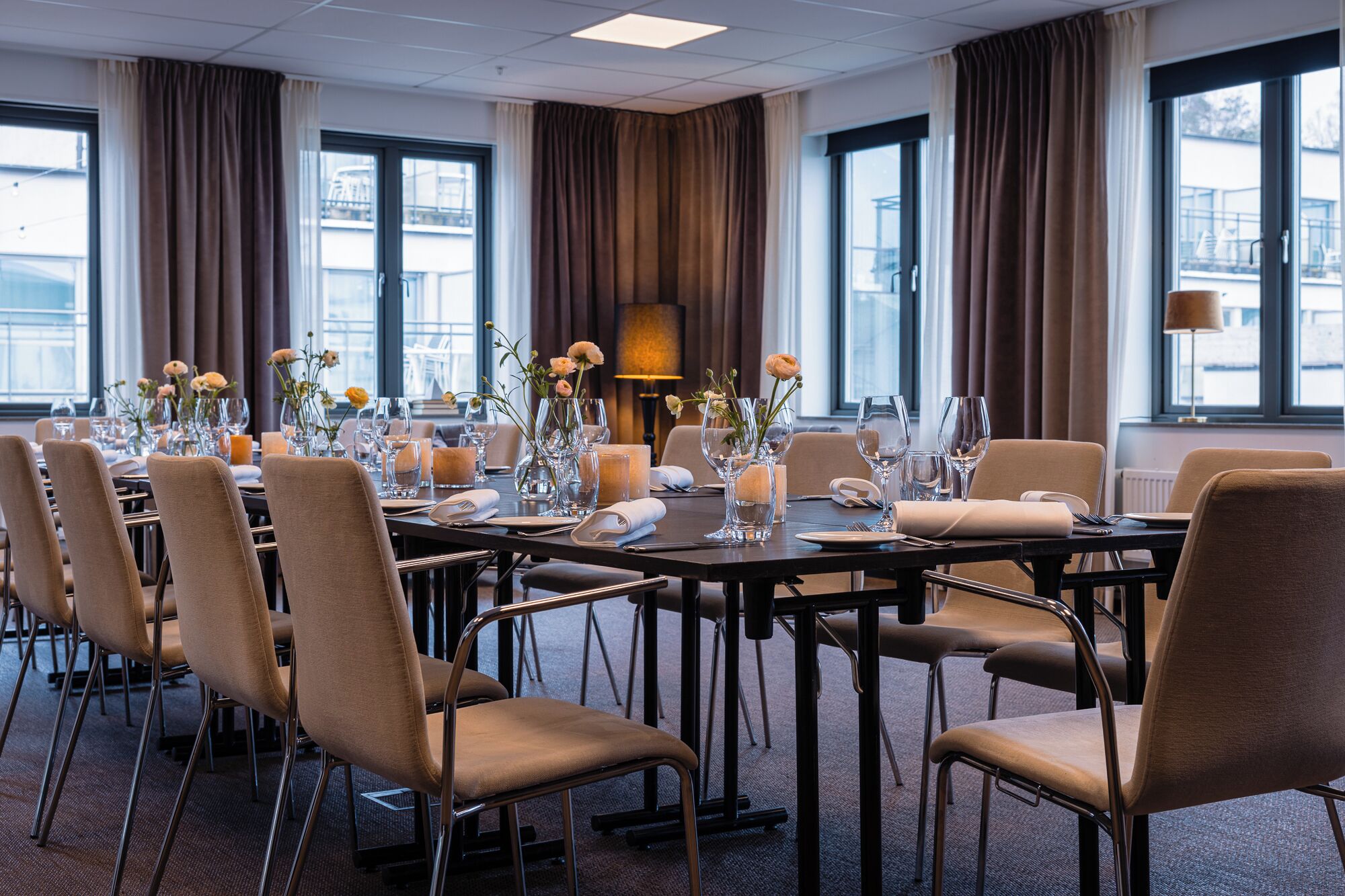 Un salón de conferencias con una larga mesa dispuesta para una reunión, equipado con sillas, vasos y centros de mesa florales.