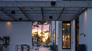 Una piscina con escalones de acceso, un área de descanso techada con sillones, y un edificio con amplias ventanas.
