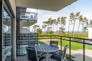 A balcony with a glass railing, a round table, and chairs, overlooking a grassy area with trees and a view of the sea.