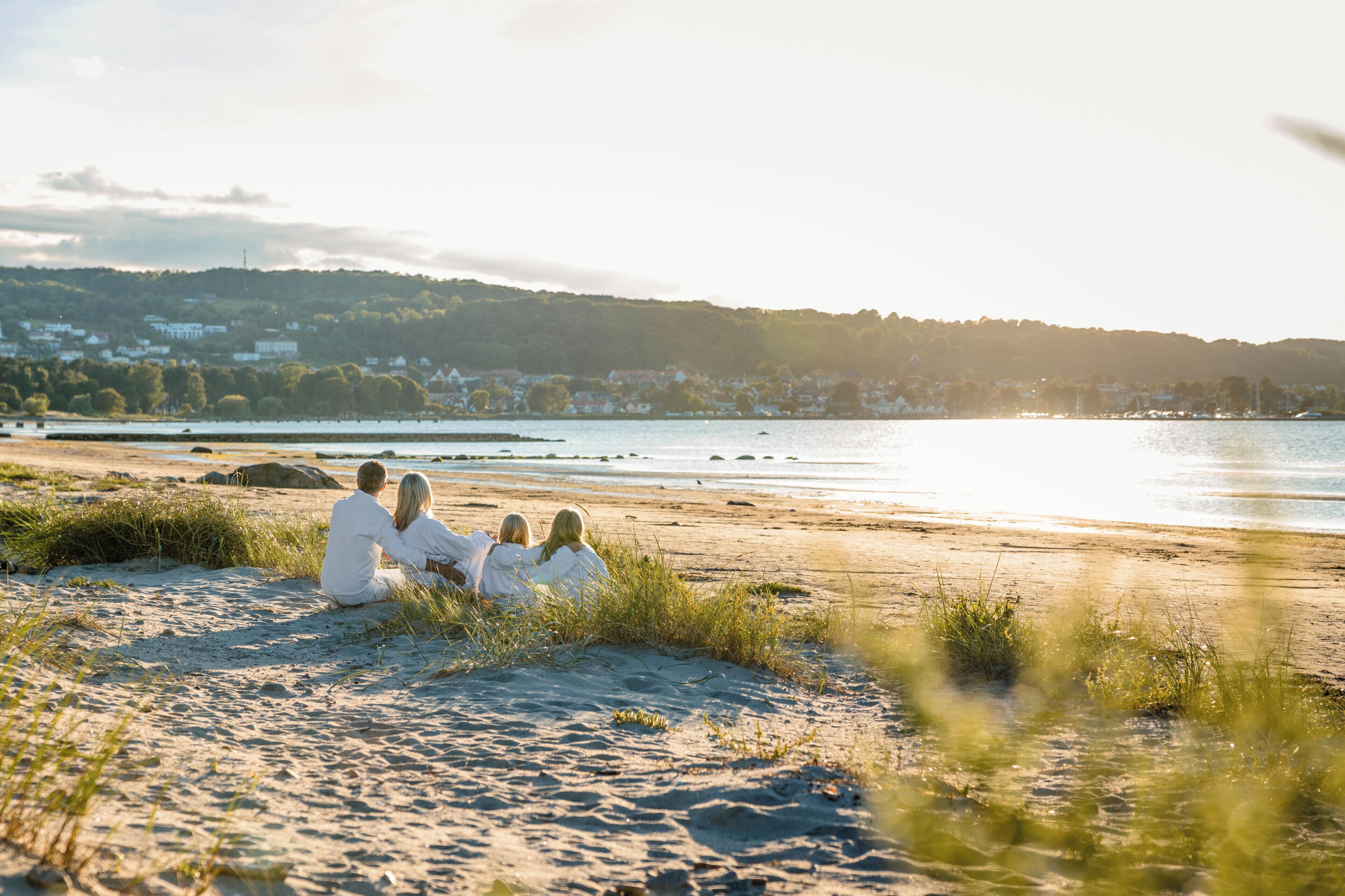 A beach with people sitting on the sand, a body of water, and a distant hill with buildings.