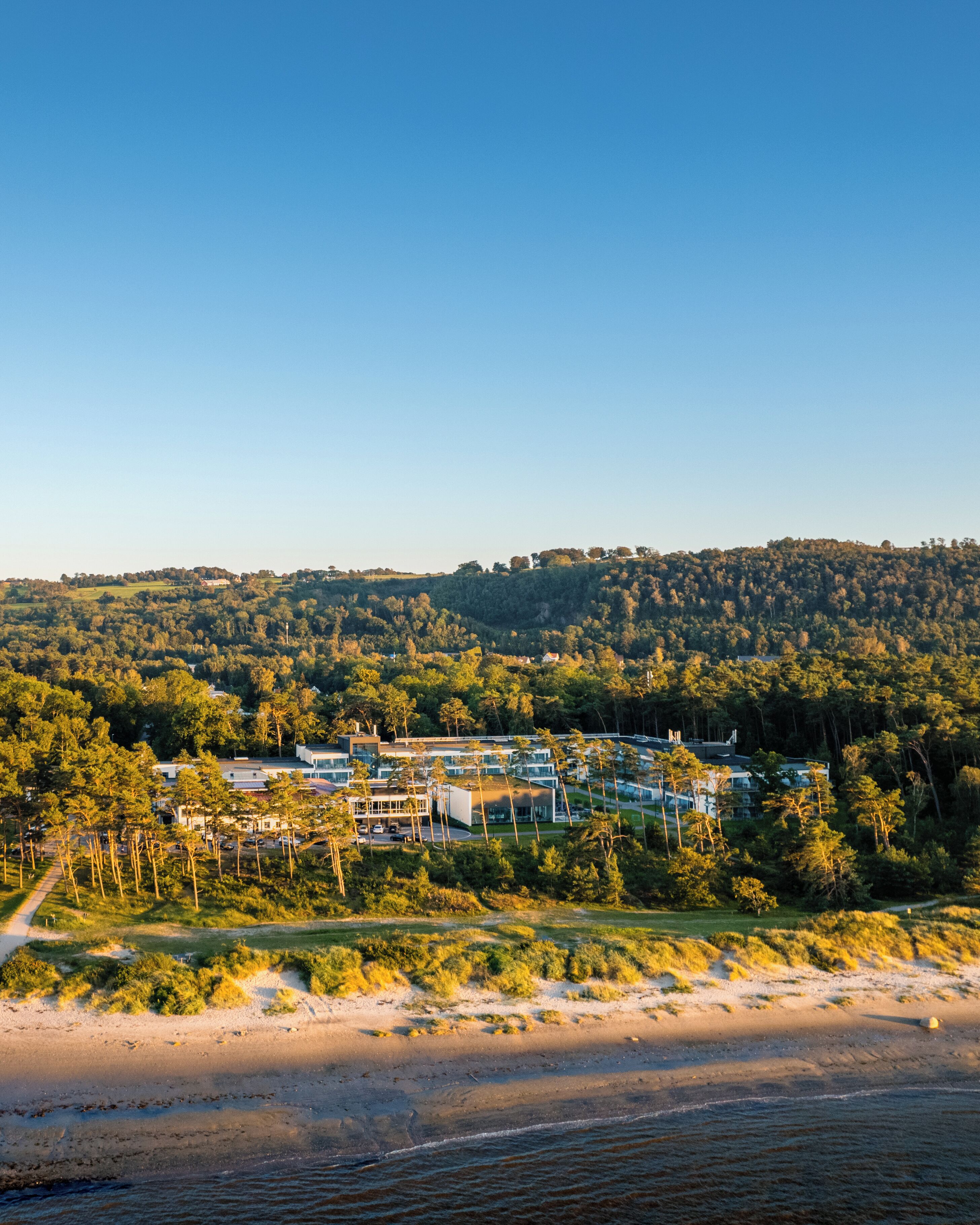 En kustort med moderna byggnader, en strand och en skogbeklädd kulle i bakgrunden.