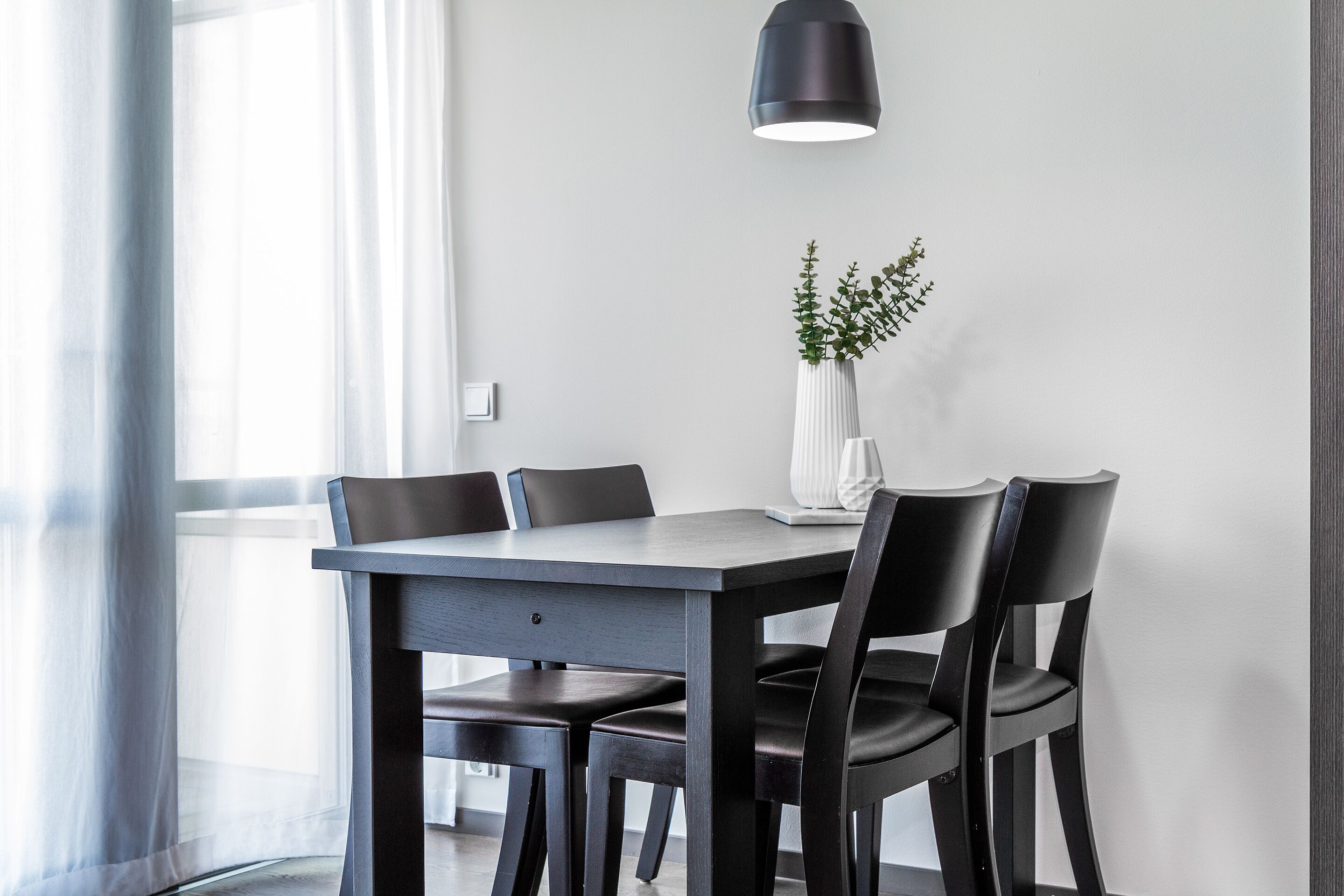 A modern dining area with a rectangular table, four chairs, a vase with green foliage, and a pendant light.