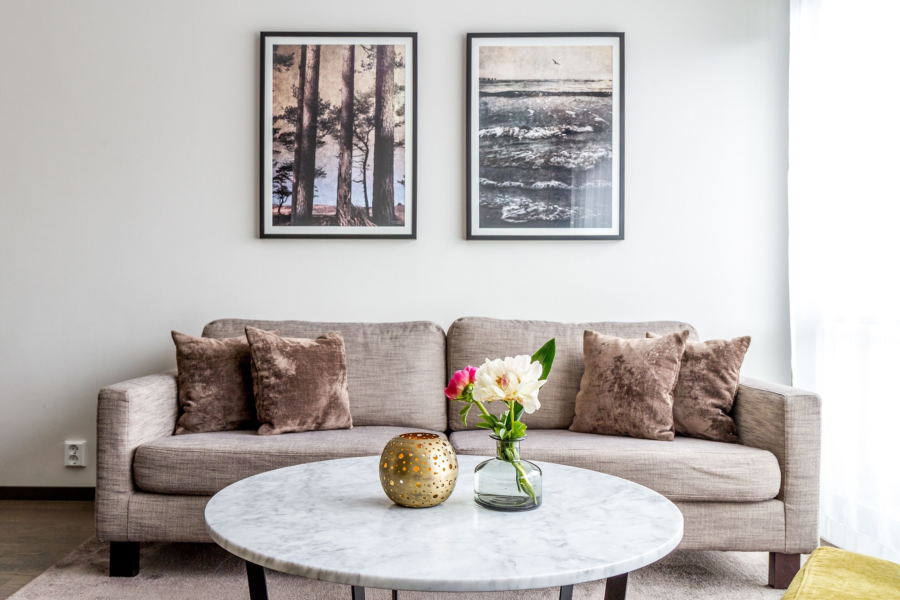 A living room with a grey sofa, two framed pictures on the wall, a round coffee table with a vase of flowers, and a decorative bowl.